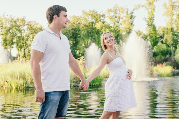 Smiling pregnant woman looking at husband near the lake in the evening holding hands. Free time outdoor.