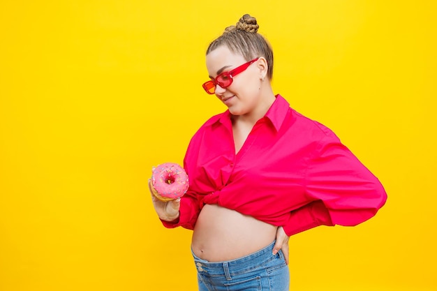 Smiling pregnant woman in pink shirt holding delicious sweet donuts on isolated yellow background Sweet caloric food during pregnancy A pregnant woman eats fatty pastries