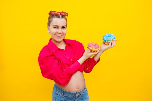 A smiling pregnant woman in a pink shirt holds donuts in her hands on a yellow background Sweet food during pregnancy Harmful food during pregnancy