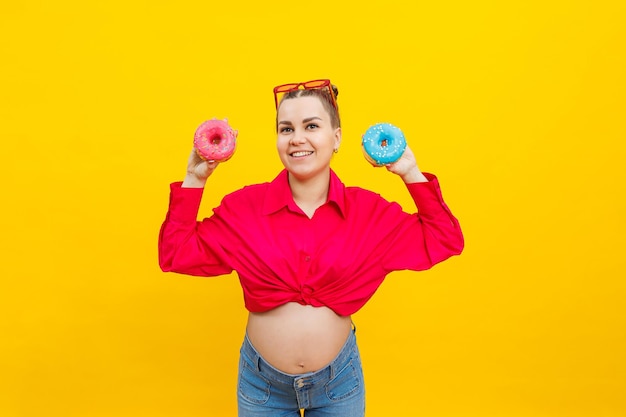 A smiling pregnant woman in a pink shirt holds donuts in her hands on a yellow background Sweet food during pregnancy Harmful food during pregnancy