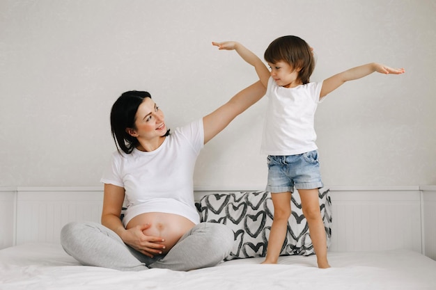 Smiling pregnant woman relaxing and playing with her little cute daughter on bed