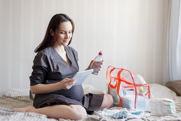 Smiling pregnant woman sitting on the bed with a list in his hands, collects things in a bag for the hospital