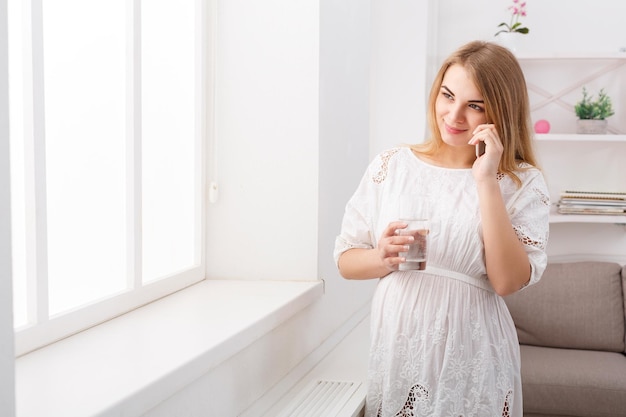 Smiling pregnant woman talking on her smartphone, copy space. Happy expectant blonde with glass of water have pleasant talk on phone.