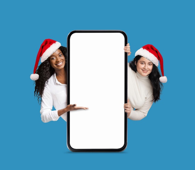 Smiling pretty african american and european millennial women in Christmas hats show hand to big phone