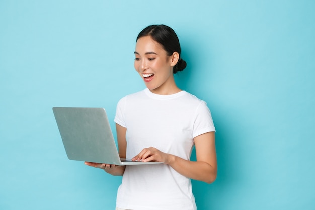 Smiling pretty asian girl in white t-shirt, working on project, looking satisfied and happy at laptop screen, freelancing or browing internet, online shopping over light blue wall