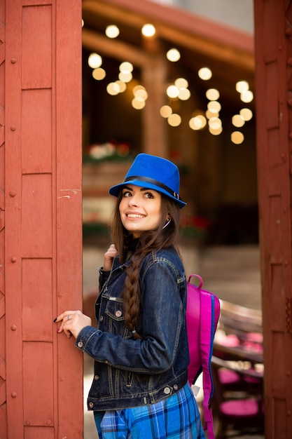 Smiling pretty girl in blue hat near old building with antique red doors. Female model posing