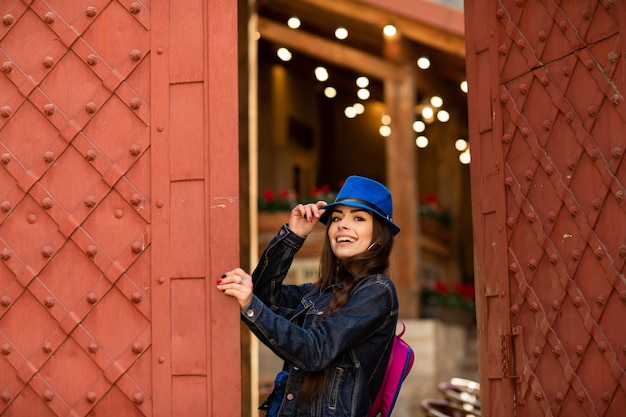 Smiling pretty girl in blue hat near old building with antique red doors. Female model posing