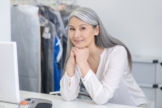 Smiling pretty gray-haired woman in white blouse standing behind dry cleaning counter waiting