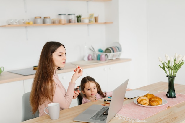 Smiling pretty woman doing make up while sitting with her little daughter at the kitchen during breakfast.