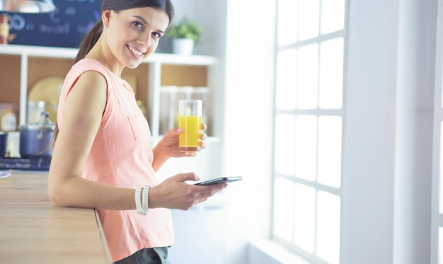 Smiling pretty woman looking at mobile phone and holding glass of orange juice while having breakfast in a kitchen