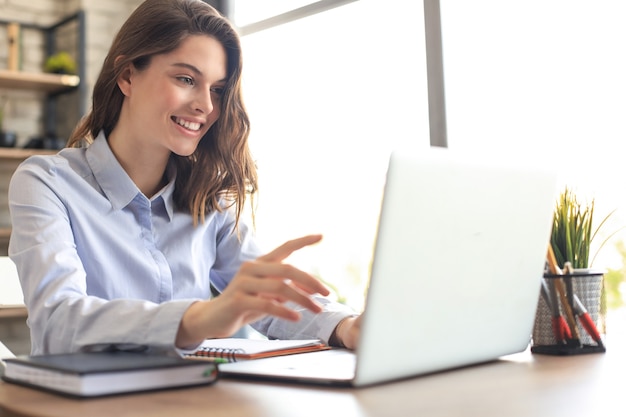 Smiling pretty woman sitting at table, looking at laptop screen. Happy entrepreneur reading message email with good news, chatting with clients online.