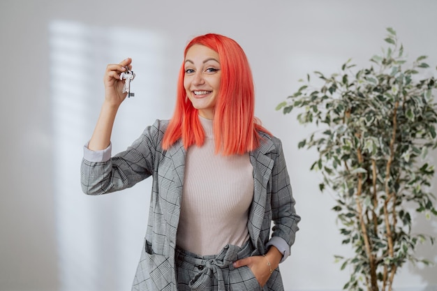 A smiling pretty woman with pink hair dressed in a smart plaid suit holds the keys to an apartment i