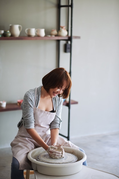 Smiling pretty young asian woman making clay vase or flower pot in pottery school