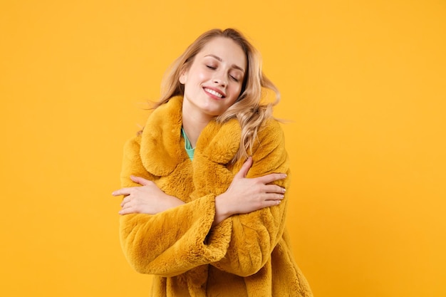 Smiling pretty young blonde woman girl in yellow fur coat posing isolated on orange background, studio portrait. People lifestyle concept. Mock up copy space. Keeping eyes closed, hugging herself.