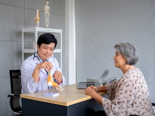 Smiling professional Asian man orthopedic doctor pointing to foot skeleton ankle joint anatomy model to explain for senior female patient in medical office Physio treatment health care concept