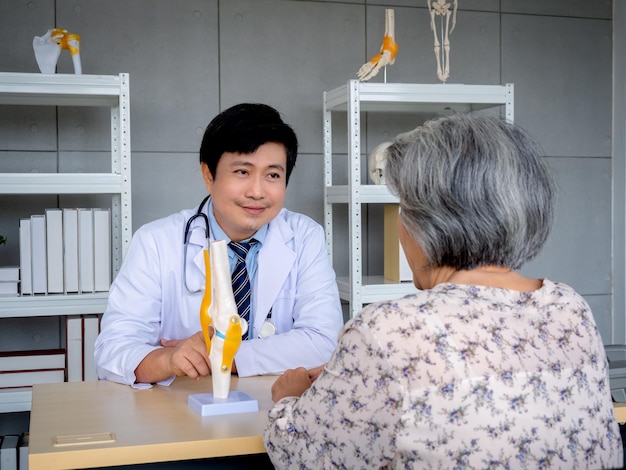 Smiling professional Asian man orthopedic doctor in white suit pointing to knee joint anatomy model to explain for senior female patient in medical office Physio treatment health care concept