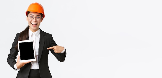 Smiling professional female construction manager asian engineer in safety helmet and business suit show project pointing finger at digital tablet screen with pleased expression white background