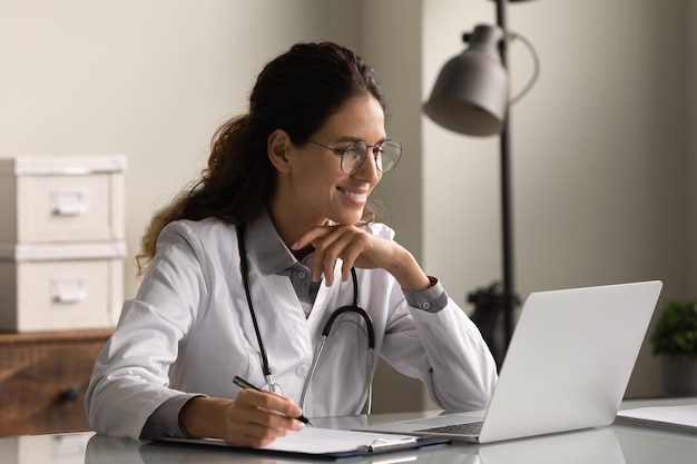 Smiling professional female doctor wearing glasses and uniform taking notes in medical journal filling documents patient illness history looking at laptop screen student watching webinar