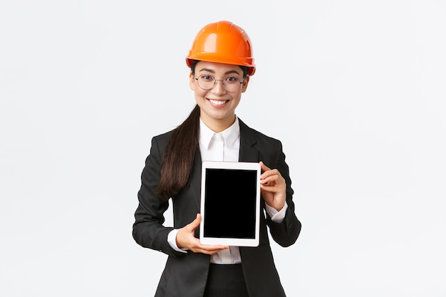 Smiling professional female engineer, saleswoman or estate agent showing location to client, standing in safety helmet and business suit, introduce construction project at tablet screen