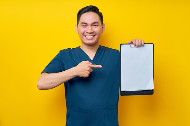 Photo smiling professional young asian male doctor or nurse wearing a blue uniform pointing finger at clipboard with blank paper isolated on yellow background healthcare medicine concept