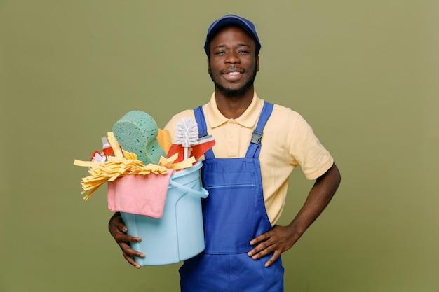 Smiling putting hand on hip holding bucket of cleaning tools young africanamerican cleaner male in uniform with gloves isolated on green background