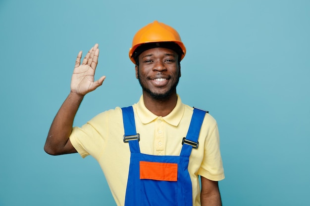 Smiling raised hand young african american builder in uniform isolated on blue background