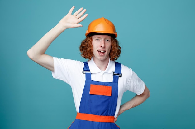 Smiling raising and putting hands on hip young builder man in uniform isolated on blue background