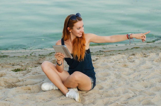 Smiling redhead slim woman sitting on a beach and holding tablet computer.