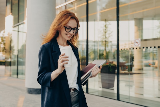 Smiling redhead woman corporate worker dressed in formal clothes drinks coffee