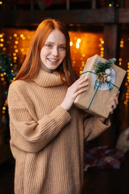 Smiling redhead young woman holding beautiful Christmas gift box on background of blurry lights