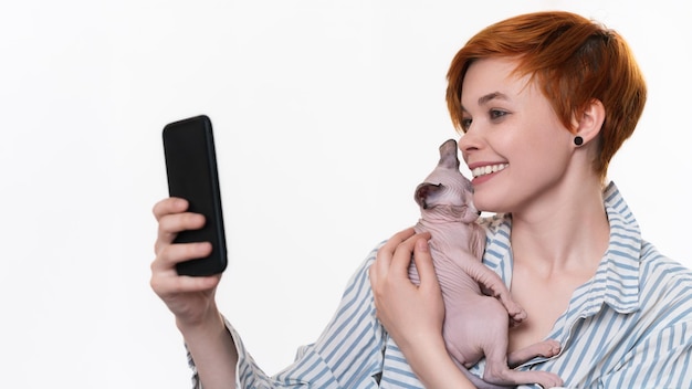 Smiling redhead young woman hugging Sphynx Cat takes selfie photo on smartphone