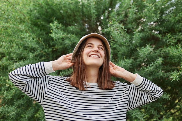 Smiling relaxed woman wearing striped shirt and panama standing with raised arms near green bush in park enjoying summertime and walking outside good weather