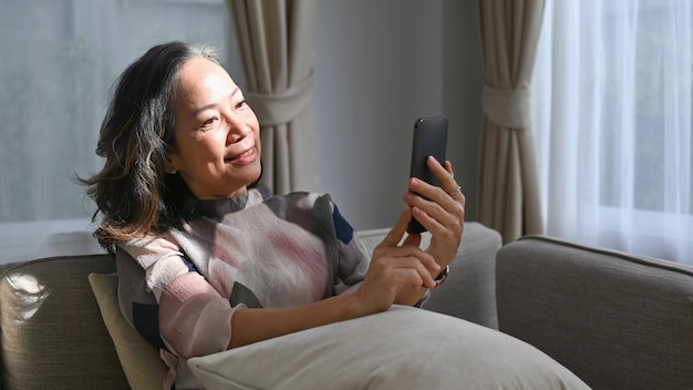 Smiling retired woman using smartphone on couch at home Retirement technology concept