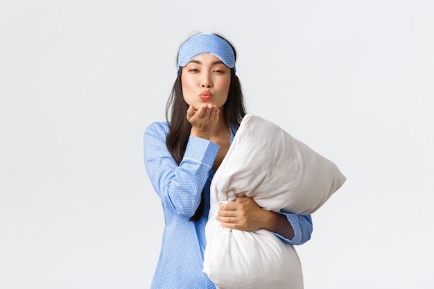 Smiling romantic asian girl in blue pajama and sleeping mask, hugging pillow and sending goodnight air kiss at camera with coquettish, sensual face, standing white background.