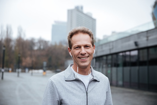 Smiling Runner in gray sportswear and headphone standing on the street and looking at camera