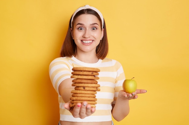 Smiling satisfied amazed woman wearing striped shirt and hair band holding out cookies and apple giving you carbs dessert instead of a healthy diet posing isolated over yellow background