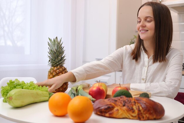 Smiling satisfied Caucasian brown haired woman wearing white shirt sitting at table in kitchen enjoying nutritious breakfast with organic fruits and vegetables healthy lifestyle