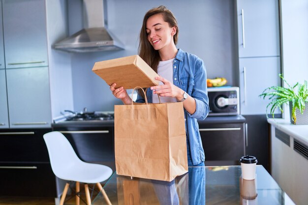 Smiling satisfied happy casual adult joyful young millennial girl customer received cardboard bags with takeaway food and drinks at home. Fast delivery service concept