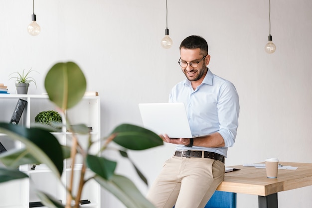 Smiling satisfied man 30s wearing white shirt sitting on table in office, and having business chat on silver laptop