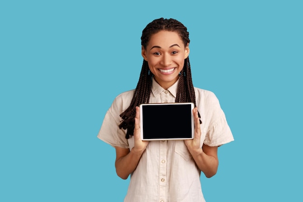 Smiling satisfied woman holds modern tablet with empty screen for your advertisement poses with electronic gadget in hands wearing white shirt Indoor studio shot isolated on blue background