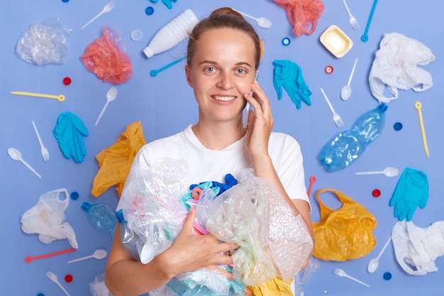 Smiling satisfied woman wearing white shirt isolated on blue wall with rubbish around holding litter talking on mobile phone looking at camera saving earth cleaning up planet