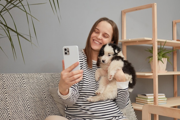 Smiling satisfied young adult woman wearing striped shirt taking selfie on smartphone with her small