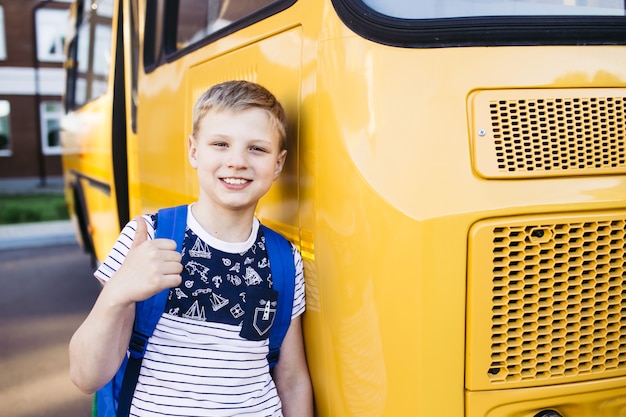 Smiling School Boy from elementary school shows a thumbs up in front of school bus. Back to school. Education. Day of knowledge.