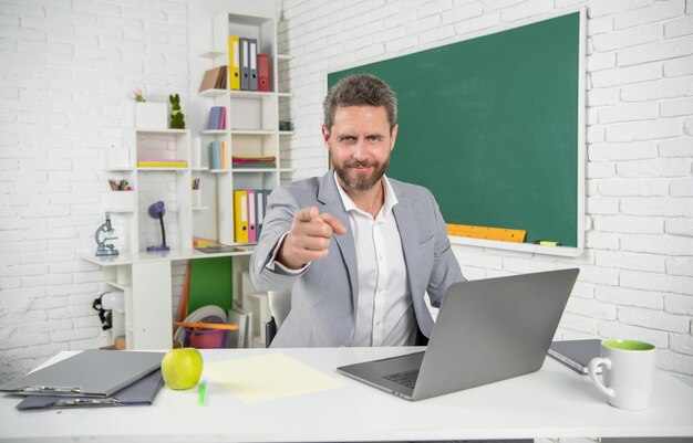 Smiling school teacher in classroom with computer at blackboard pointing finger