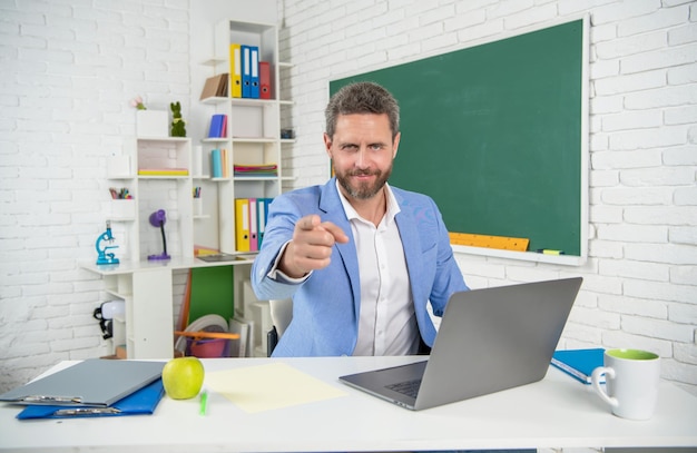 Photo smiling school teacher in classroom with computer at blackboard pointing finger