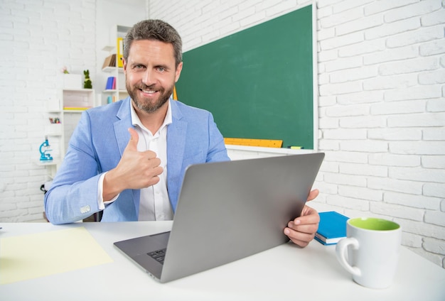 Smiling school teacher in classroom with computer at blackboard