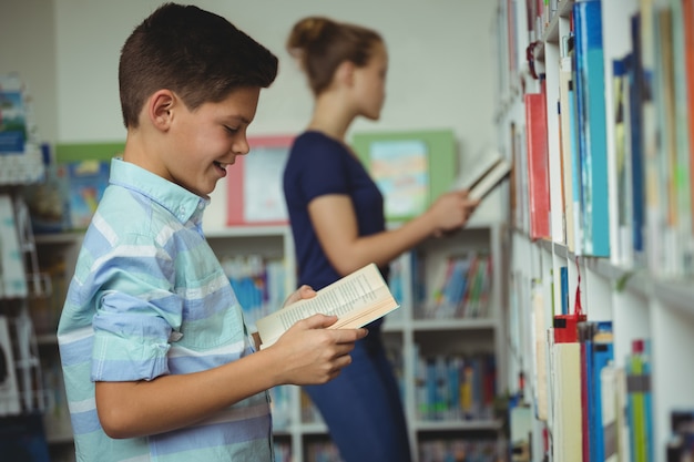 Smiling schoolboy reading book in library