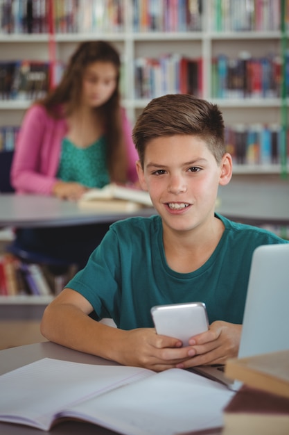 Smiling schoolboy using mobile phone in library at school