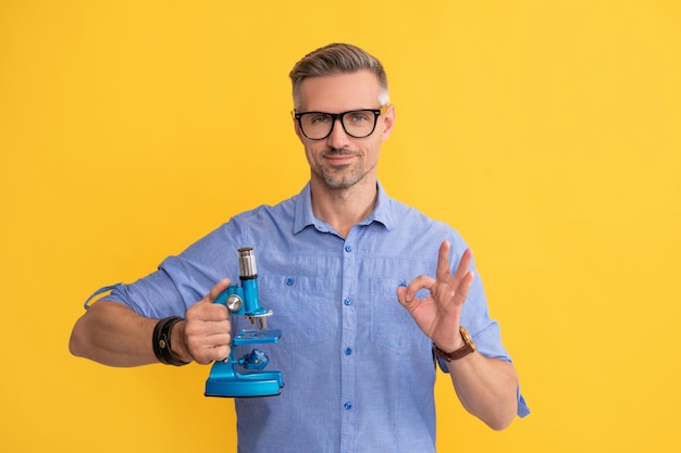 Smiling scientist man in glasses hold microscope show ok gesture on yellow background science