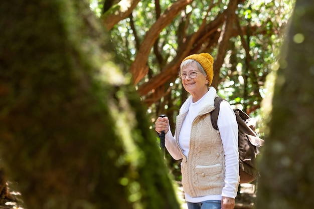 Smiling senior caucasian woman with backpack and cap hiking in the forest with walking stick enjoying nature and healthy lifestyle